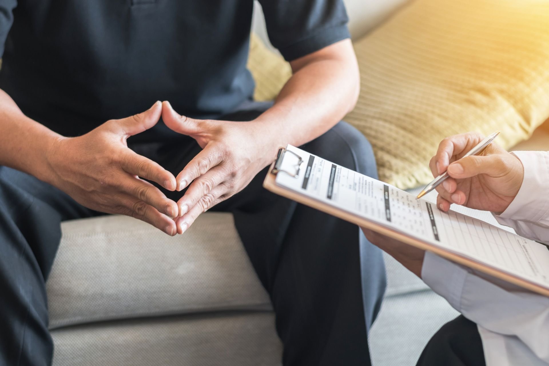 a man sitting on a couch holding his hands together and another man/woman holding a clipboard with a form